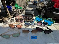 Photo showing ten assorted sunglasses and eyeglasses on a table outdoors, includes Guess branded case and soft pouch, two novelty green cloud shaped blue lens sunglasses visible.