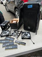 Thermaltake PC case with side door open showing accessories including RAM sticks, fans, and mounting brackets on white table.