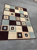 Image showing full rectangular rug with pattern of squares and rectangles in various earth tones and muted blue, laid on pavement for display.