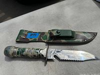 Full view of serrated survival knife lying above camouflage sheath on a flat surface showing blade and handle details.