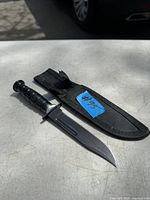 Photo of a fixed blade knife with a black grooved handle lying next to its black fabric sheath case on a white surface outside.