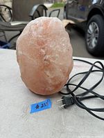 Photo shows a natural pink Himalayan salt lamp with an electrical cord and plug on an outdoor table with chairs and car in background.