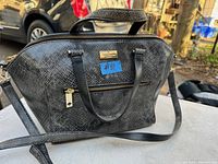 Front view of a black faux snake skin Kate Spade purse with zippered front pocket, dual handles, and shoulder strap on an outdoor table.