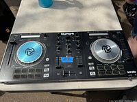 Numark MT Pro3 DJ mixing board laid flat on a white surface outdoors, showing dual silver-blue jog wheels, central mixer controls, and button pads on either side.