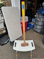 Sledge hammer standing vertically on a white plastic bench in a garage setting with various items in the background.