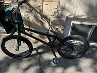 Side view of black BMX bicycle showing the full frame, tires, blue pedals, and gray seat against a stone wall background.