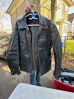 Vintage men's thick black leather jacket hanging on a plastic hanger outside, showing full front view including collar, zipper, pockets, and sleeves.