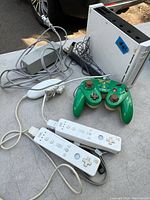 White Nintendo Wii console with two green GameCube controllers, two white Wii remotes with MotionPlus, power adapter and sensor bar cables on table.