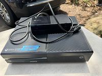 Black Xbox One console with power cord placed on a table outdoors. Shows front view with HDMI label, power button, and Xbox logo.