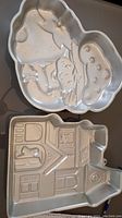 Two aluminum cake pans shown on a table. One is shaped like Strawberry Shortcake, the other shaped like a haunted house with ghost and bats.