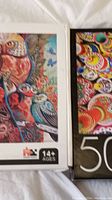 Close-up of two puzzle boxes showing colorful artwork and 500-piece count with recommended age 14+. One box features bird illustrations, the other a multicolored pattern.