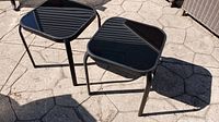 Two matching outdoor patio side tables on stone patio with black shiny tops and metal frames. One table is slightly lower than the other.