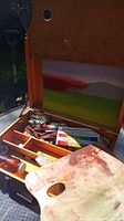 Open artist painting box showing compartments holding paint tubes, wooden palette with thumb hole, and slots for canvases with a partially visible canvas inside the lid