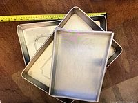 Three metal rectangular cake pans with one pan showing a dent. Visible measuring tape shows approximate pan sizes.