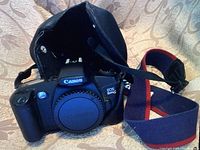 Photo of Canon EOS Rebel G 35mm film camera body with body cap, black protective case, and red/blue strap on a patterned fabric background.