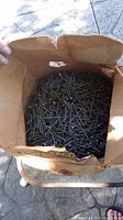 Open cardboard box with a partial quantity of metallic flat head nails inside
