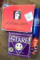 Photo shows three games: Scattergories in a worn red box, Stare! in a purple box with a smiley face design, and The World Master Cube puzzle in a tall blue tube with orange and white details.