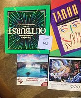 Four boxed items: Outburst board game in green box, Taboo board game, 550-piece Niagara Falls postcard collection puzzle, 1000-piece fairy scene glitter puzzle.