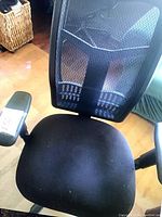 Front view of black office chair with mesh backrest and cushioned seat, showing adjustable armrests with control buttons, situated indoors on a wood floor.