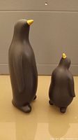Front view of two black penguin figurines with yellow beaks, one larger and one smaller, standing upright on a beige surface.
