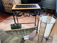 Photo showing a small metal-leg table with wooden lattice top, two decorative wall plaques, and a small cylindrical lamp with fabric shade.