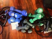 Photo shows a collection of blue, green, and gray hand weights on a wooden floor along with multiple resistance bands with black handles.