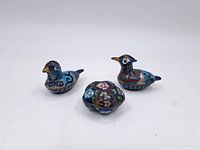 Three enameled brass trinket boxes: two bird-shaped with detailed blue, red, black, white, and gold enamel patterns, and one flower-shaped box with multicolored floral enamel decoration, all placed on a white surface.