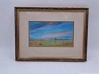Frontal photo of framed watercolor landscape showing field, sky, and distant buildings, with signature visible.