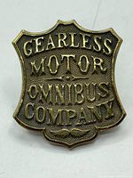 Front view of a shield-shaped brass staff cap badge with embossed text 'GEARLESS MOTOR OMNIBUS COMPANY'.