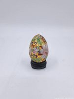 Front angle view of the small cloisonne egg showing floral and bird enamel patterns on a colorful gold-tone background mounted on a black stand.