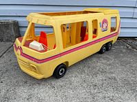 Side front view of yellow Barbie motorhome toy showing red seats inside and wheels, placed on concrete ground