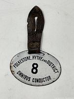 Front view of oval white enamel badge with 'FOLKESTONE, HYTHE AND DISTRICT 8 OMNIBUS CONDUCTOR' in black lettering, attached to worn leather strap.