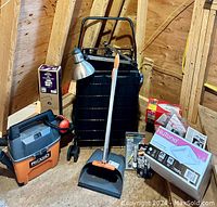 Photo showing oversized black metal shopping cart with removable canvas sack, Rigid wet/dry vacuum, desk lamp, dust pan and broom, unopened NuTone fan box, and miscellaneous small items grouped together on attic floor.