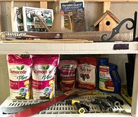 Front view of gardening supplies including plant food bags, burlap rolls, machetes, bird house, hose nozzles, and gardening tools arranged on white shelving.