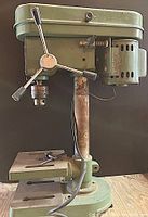 Front and side view of Ludell DP-3 drill press showing three-spoked feed lever, motor, chuck, and base with clamping slots.