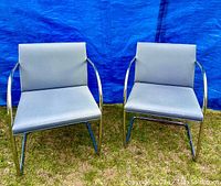 Front view of both chairs showing blue-gray upholstery and shiny tubular steel frames on grass background with blue tarp behind.