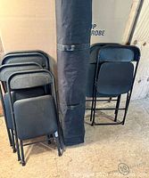 Photo showing six black vinyl folding chairs stacked and the tent case standing upright behind them.