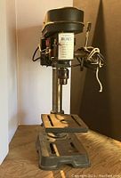 Front view of older 5-speed drill press on wooden surface, showing base, adjustable work table, and head with motor housing.