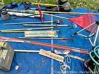 Photo showing assorted gardening tools including rakes, hoes, rolling seeders, shovels, sprinkler heads and wooden rods spread on a blue tarp outdoors.