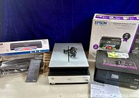Photo showing Epson Expression Premium XP-6000 printer in box, Epson Perfection V700 scanner with cords on top, and one other item possibly related to the laminator parts out of box.