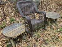 Dark brown resin wicker rocker chair centered with two small rustic wooden tables on each side, all placed on leaf-covered ground with stone background