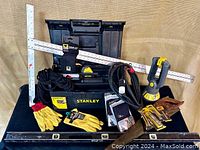 Full view of Stanley 19" black plastic toolbox with tools and accessories including gloves, rope, clamp flashlight, T-square and more as arranged on backdrop.