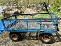 Side view of large blue steel garden utility wagon with mesh sides and pneumatic tires on rough terrain.