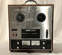 Front view of TEAC A-2070 reel-to-reel tape deck showing the two reel spindles, control knobs, and VU meters in wooden casing.