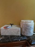 Two marble keepsake boxes on a dark marble surface, a rectangular box with black handle on the left, a cylindrical box on the right