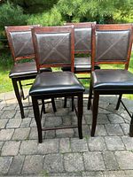 Front view of all four wooden tall chairs with dark brown cushioned seats and backs, arranged outdoors on stone pavement.