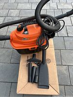 Overview of the Orange and black portable Armor All car vacuum, with a flexible hose and multiple black plastic attachments arranged on a cardboard box.