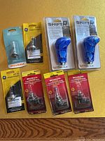Overview of halogen bulbs including Sylvania, GE H7-55 bulbs and MetraMate 9005, 9006 bulbs, plus two blue SHIFT-R shifter knobs in original packaging all laid out on yellow surface.