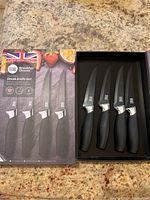 Two brand new 4-piece steak knife sets in original packaging, each with black handles and serrated blades coated with ceramic.