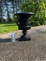 Full view of the black cast iron urn planter standing outdoors next to a water bottle for size scale.
